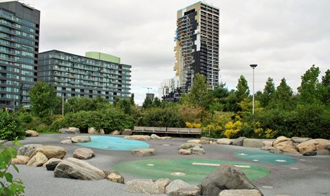 safety surface play area with embedded natural boulders. Benches at far side with trees and shrubs behind. Tall apartment buildings in background